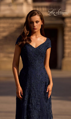 A woman stands outdoors in a navy blue Long Formal Dress: La Femme 27915, with a blurred stone building behind and the “La Femme” logo in the upper right corner.