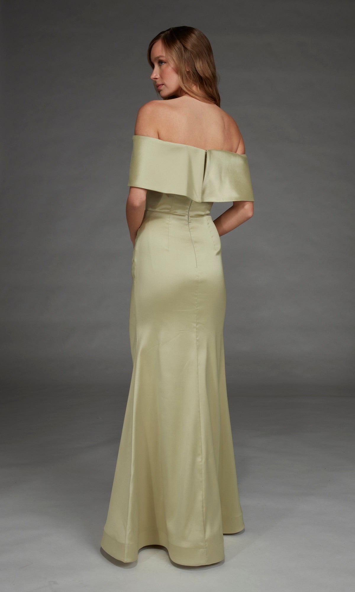 A woman stands with her back to the camera against a plain studio backdrop, wearing the Alyce 27916—a floor-length, off-the-shoulder long formal dress in light gold stretch mikado.