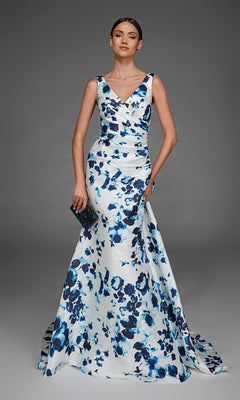 A woman wearing the Alyce 27917 long formal dress in blue/white, a sleeveless floor-length v-neck evening gown with floral patterns, holds a small black clutch and stands against a plain gray background.