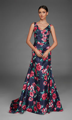 A woman wearing the Alyce 27917 long formal dress in Midnight—a floor-length, sleeveless V-neck evening gown featuring a bold red and pink floral pattern on a dark background—stands against a plain gray backdrop.