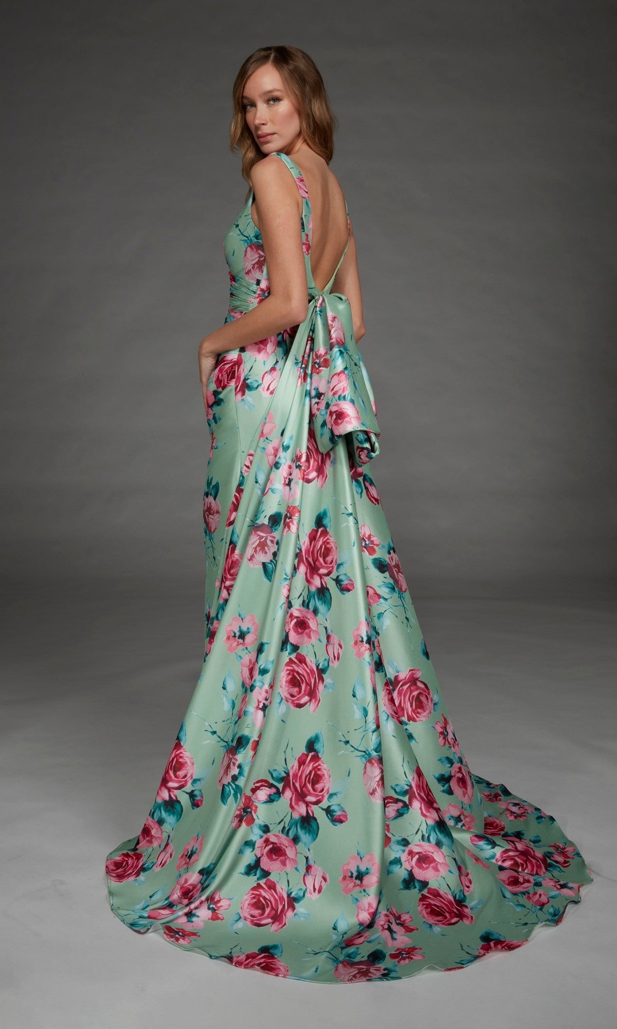 A woman wearing the Alyce 27917 long formal dress—a sleeveless, floor-length mint green evening gown featuring a v-neckline, pink floral print, and dramatic open back—stands against a gray background.