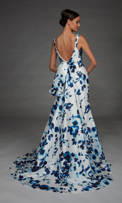 A woman wearing the Long Formal Dress: Alyce 27917—a dramatic, floor-length, backless white evening gown featuring a blue floral print and a large bow—stands against a plain gray background.