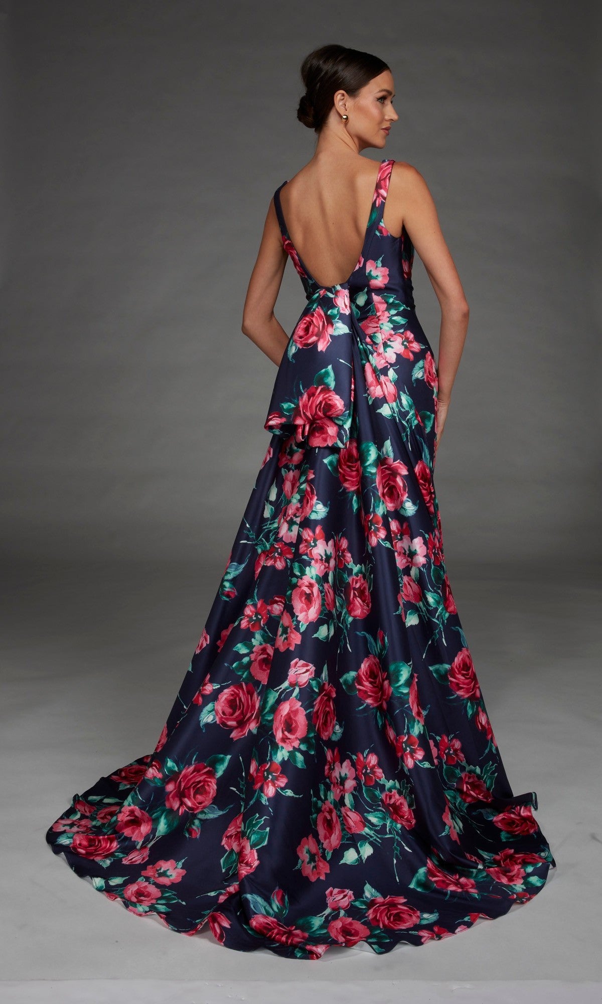 A woman poses against a plain gray background wearing the Alyce 27917 long formal dress, a sleeveless, floor-length navy gown with a v-neckline, deep V-back, pink floral print, and an elegant train.