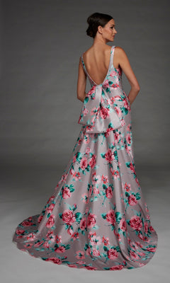 A woman wears the Alyce 27917 Long Formal Dress, a floor-length, sleeveless v-neck floral evening gown with a deep V-back and large bow, standing against a plain gray background.