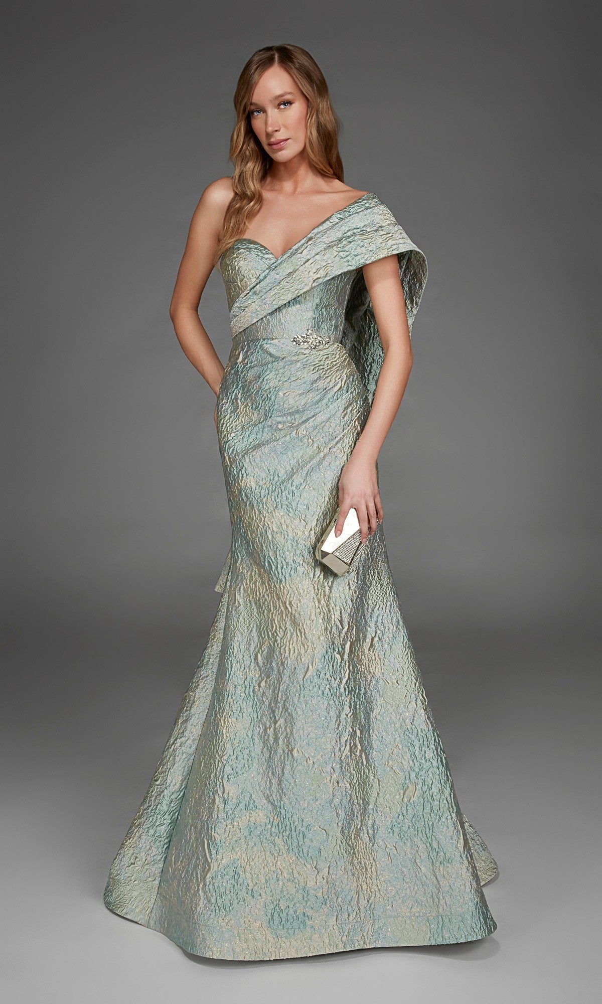 A woman stands in the Alyce 27918 long formal dress in celadon, featuring a textured design and a sweetheart neckline, holding a small clutch against a plain gray background.
