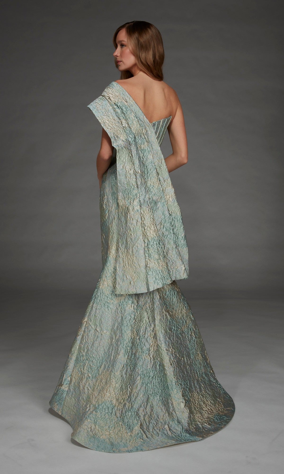 A woman stands against a plain gray background, facing away from the camera, wearing the Alyce 27918 long formal dress—a textured, metallic jacquard one-shoulder gown with draped fabric.