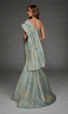 A woman stands against a plain gray background, facing away from the camera, wearing the Alyce 27918 long formal dress—a textured, metallic jacquard one-shoulder gown with draped fabric.