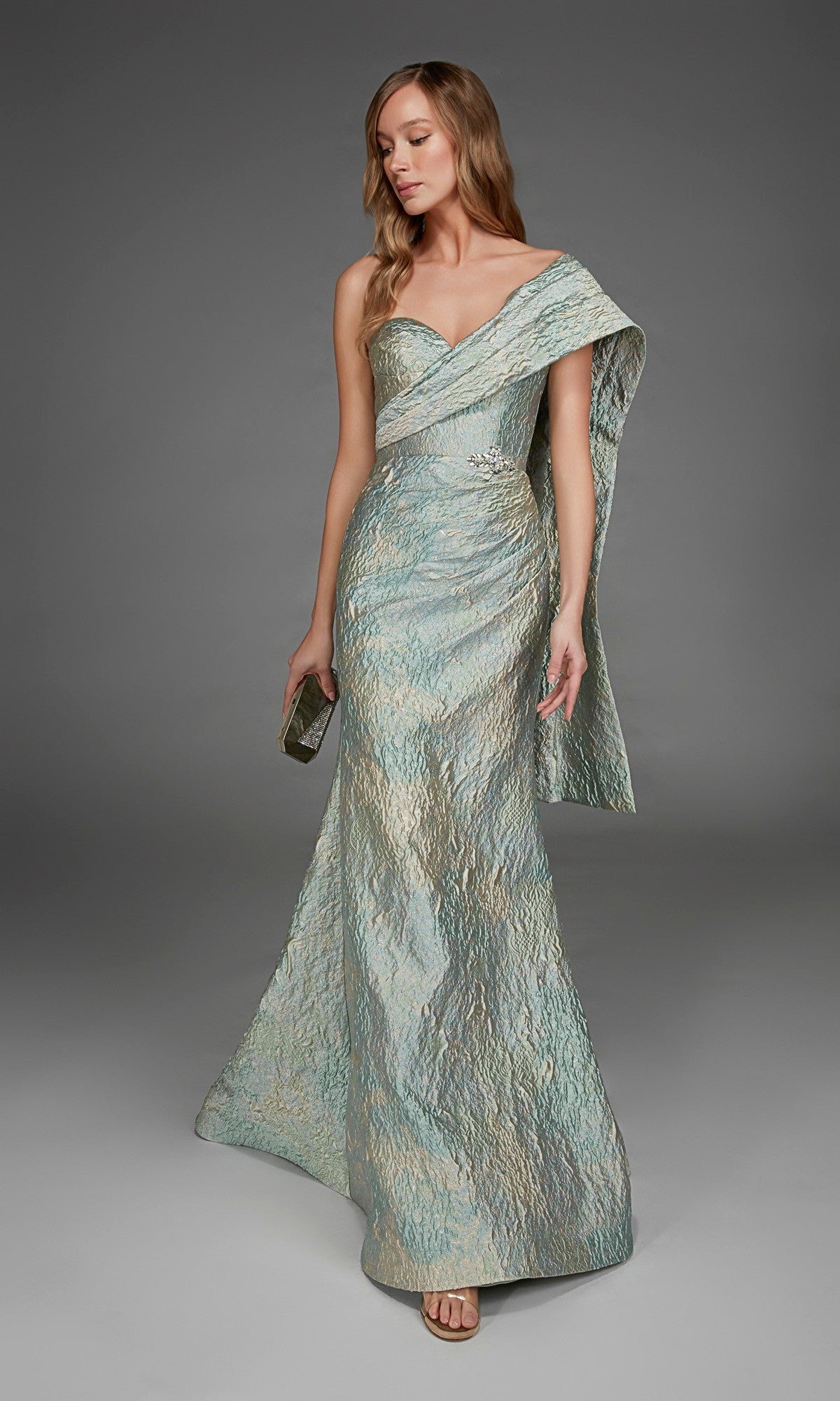 A woman wearing the Alyce 27918 long formal dress—a shimmering, metallic jacquard one-shoulder gown with a side drape—holds a clutch and stands against a plain gray background.