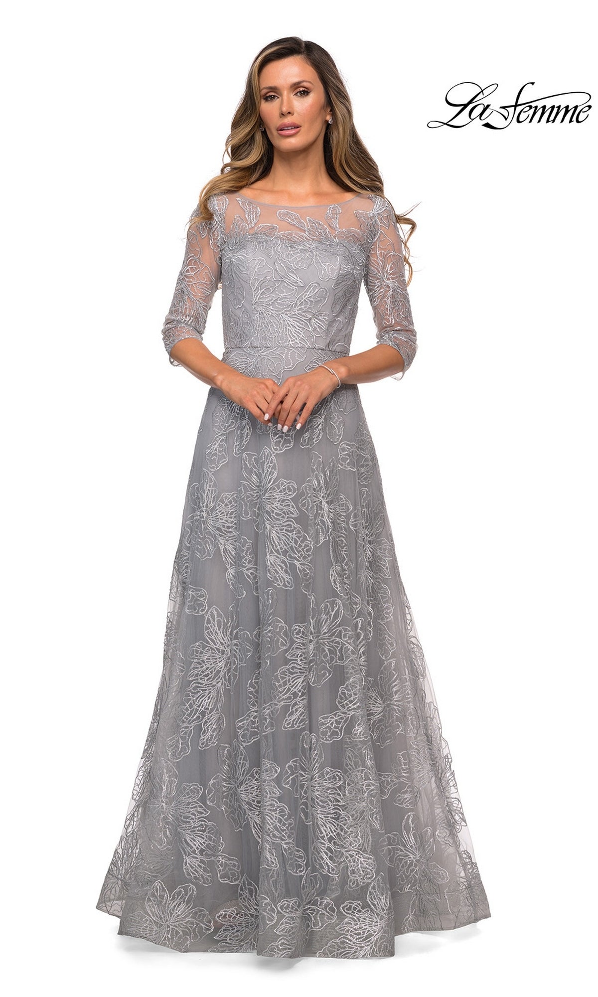A woman stands facing forward in the Long Formal Dress: La Femme 27942, a silver, modest, floor-length gown with three-quarter sleeves and floral lace details. La Femme logo top right.