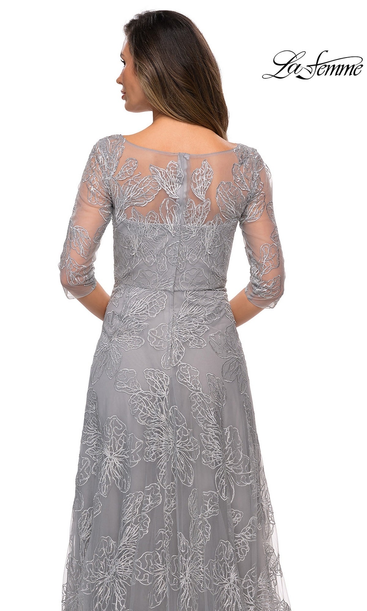 Woman in a sheer, embroidered gray Long Formal Dress: La Femme 27942 with three-quarter sleeves is shown from the back, head turned slightly. “La Femme” logo appears above.