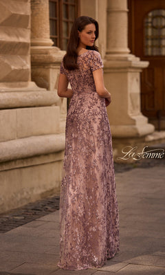 A woman wearing the Long Formal Dress: La Femme 27956 with lavender lace and floral beading stands on a cobblestone street, facing away near a stone building.