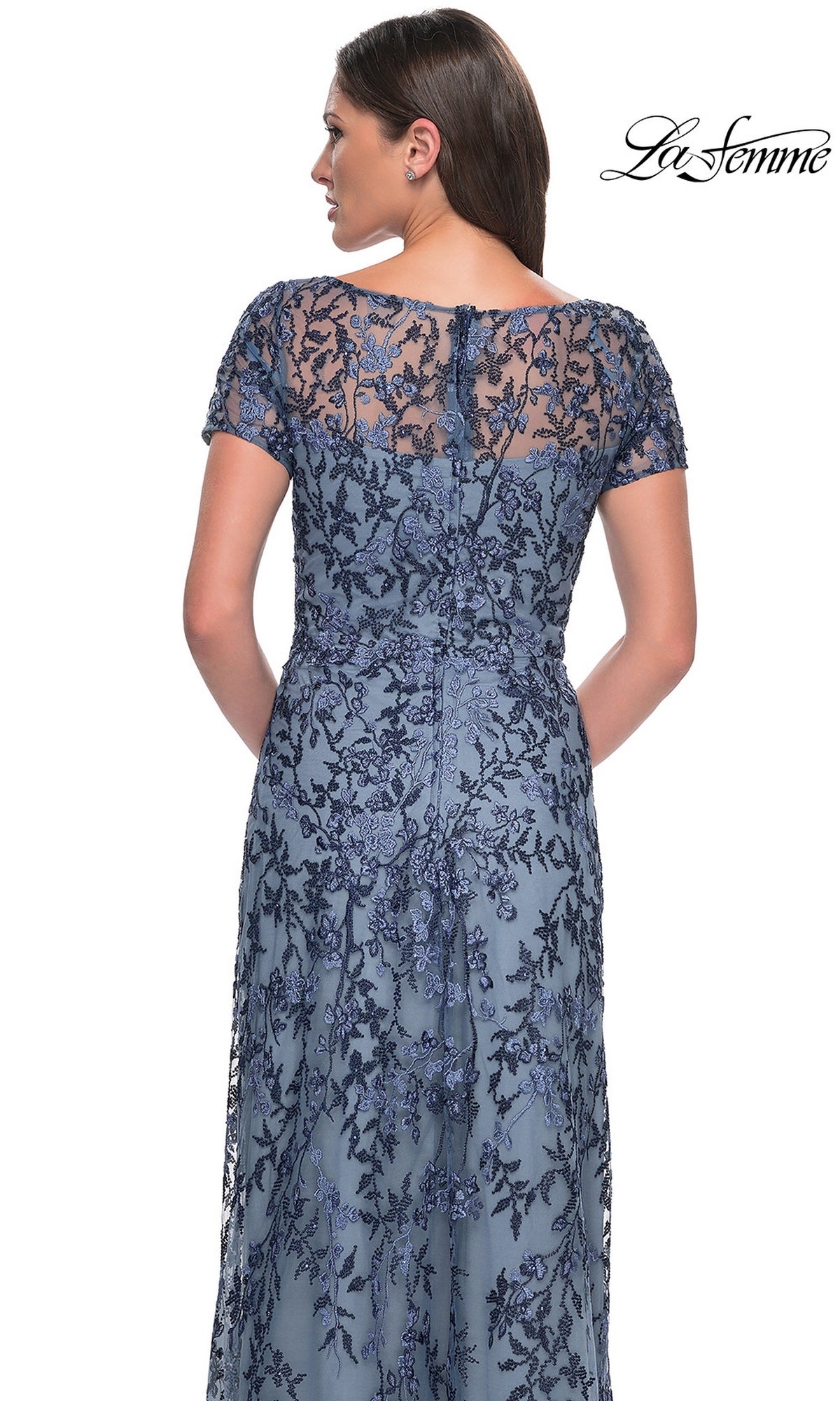A woman wears the Long Formal Dress: La Femme 27956—blue lace, floral beaded embellishments, short sleeves, and a sheer back panel. “La Femme” logo in the top right corner.