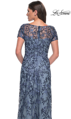 A woman wears the Long Formal Dress: La Femme 27956—blue lace, floral beaded embellishments, short sleeves, and a sheer back panel. “La Femme” logo in the top right corner.