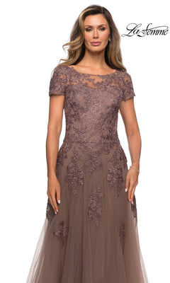 A woman wears the Long Formal Dress: La Femme 27958, a cocoa floor-length gown featuring a tulle A-line skirt, short sleeves, and a bodice adorned with floral lace embroidery.