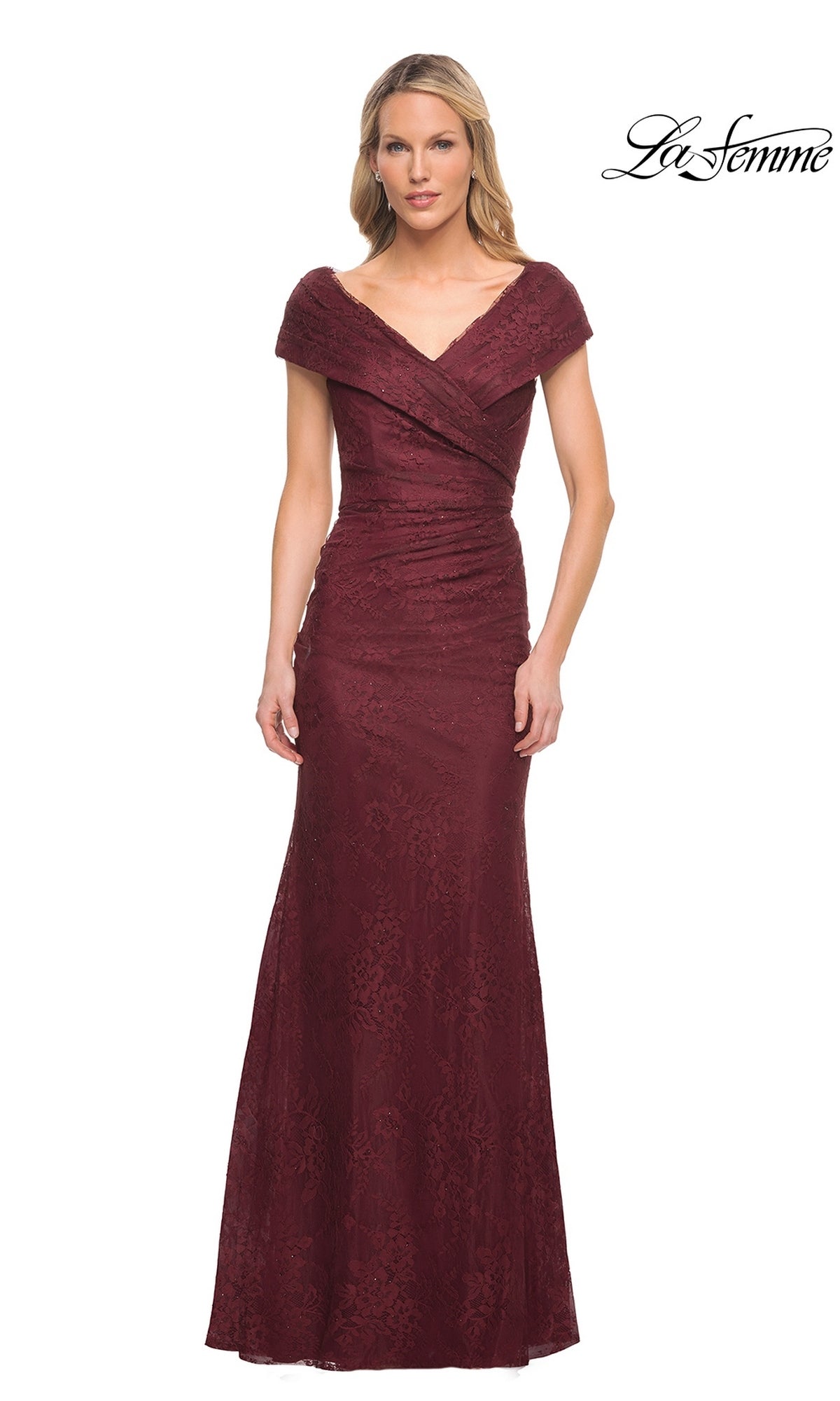 A woman stands against a white background wearing the “La Femme 27982” long formal dress in burgundy, a floor-length, off-the-shoulder mother of the bride gown with short sleeves. The elegant dress features an alluring v-neckline and delicate lace detailing. The “La Femme” logo is visible in the top right corner.