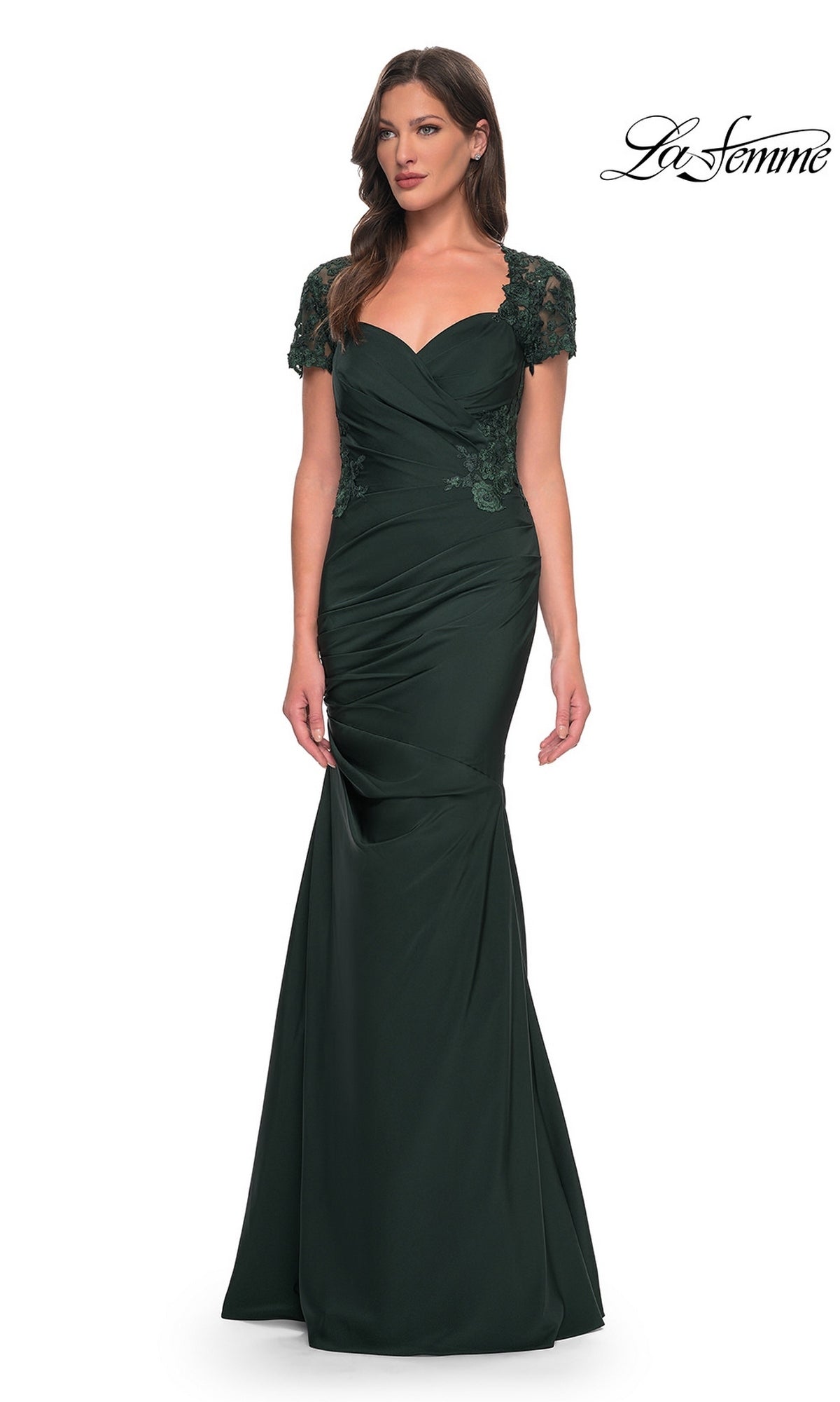A woman wearing the La Femme 27989 long formal dress in dark emerald, featuring a floor-length satin-jersey design with short lace sleeves and a ruched bodice, stands against a plain white background.