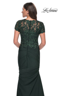 A woman is seen from behind wearing the Long Formal Dress: La Femme 27989 in dark green satin jersey, featuring lace applique detailing on the upper back and short sleeves. The elegant trumpet silhouette accentuates her figure, while the “La Femme” logo is visible in the top right corner.