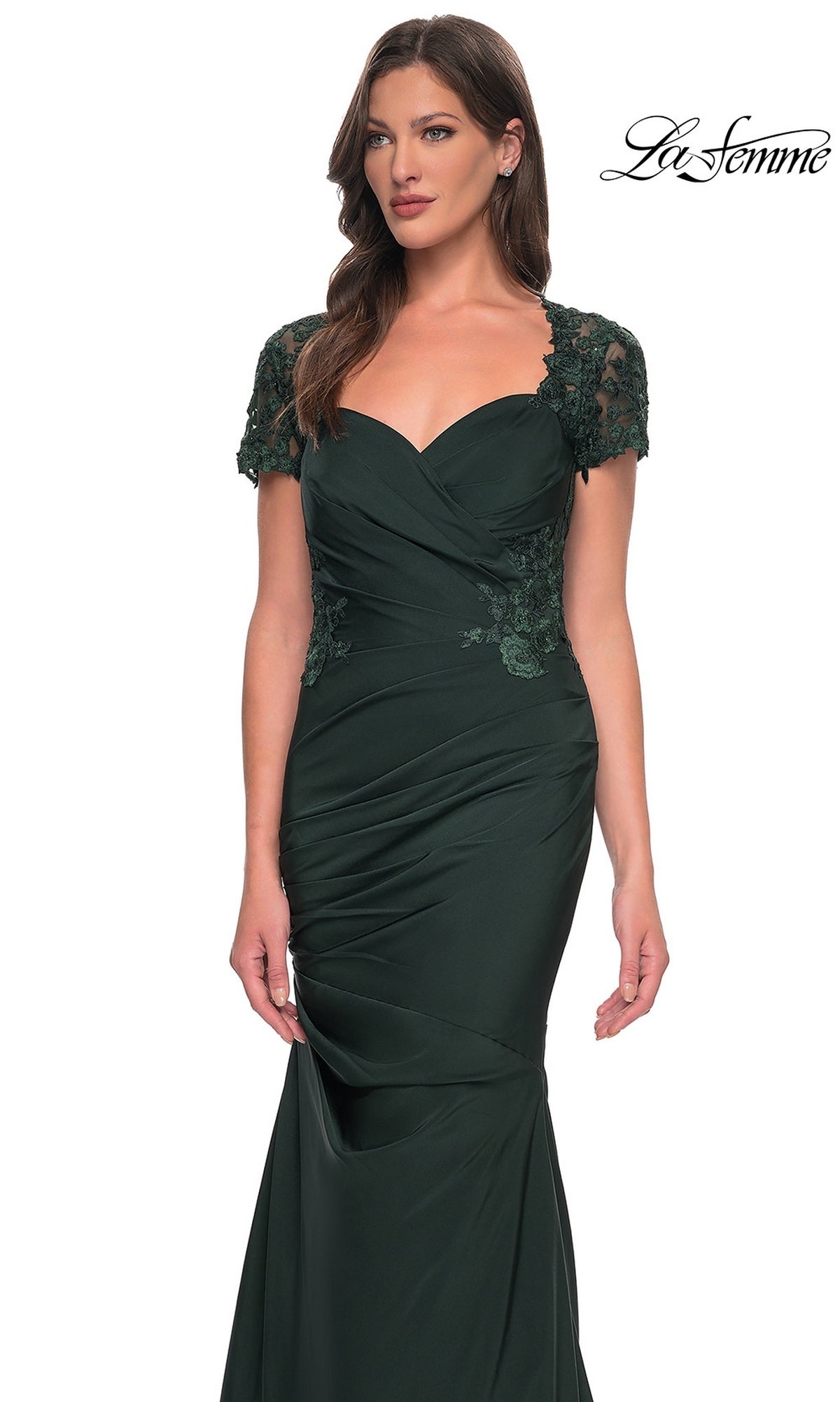 A woman wearing the La Femme 27989 Long Formal Dress— a dark green, fitted evening gown with short lace sleeves, ruched detailing, and delicate lace applique—stands against a plain white background.
