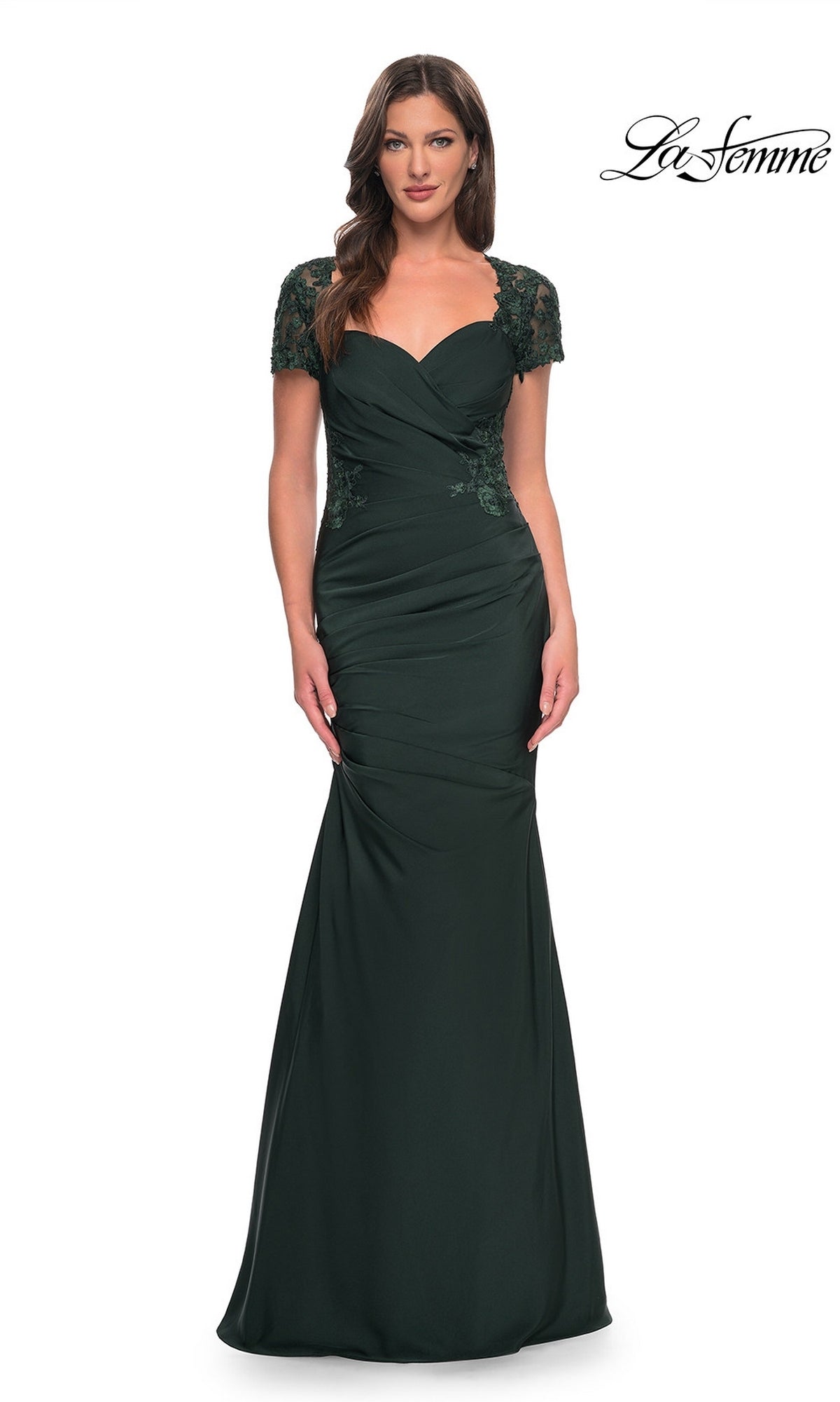 A woman stands against a white background wearing the La Femme 27989 long formal dress, featuring dark green satin-jersey fabric, a floor-length silhouette, short lace sleeves, a ruched bodice, and a sweetheart neckline. The word “La Femme” appears in the top right.