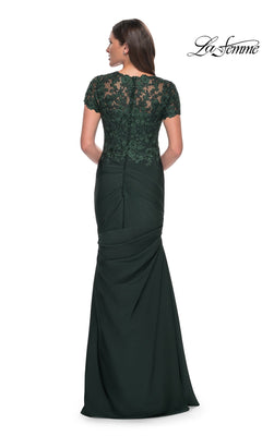 Woman in the La Femme 27989 long formal dress—a floor-length dark green gown crafted from elegant satin-jersey. The dress features a graceful trumpet silhouette and is beautifully accented with intricate lace applique on the back and shoulders, as she stands with her back facing the camera.