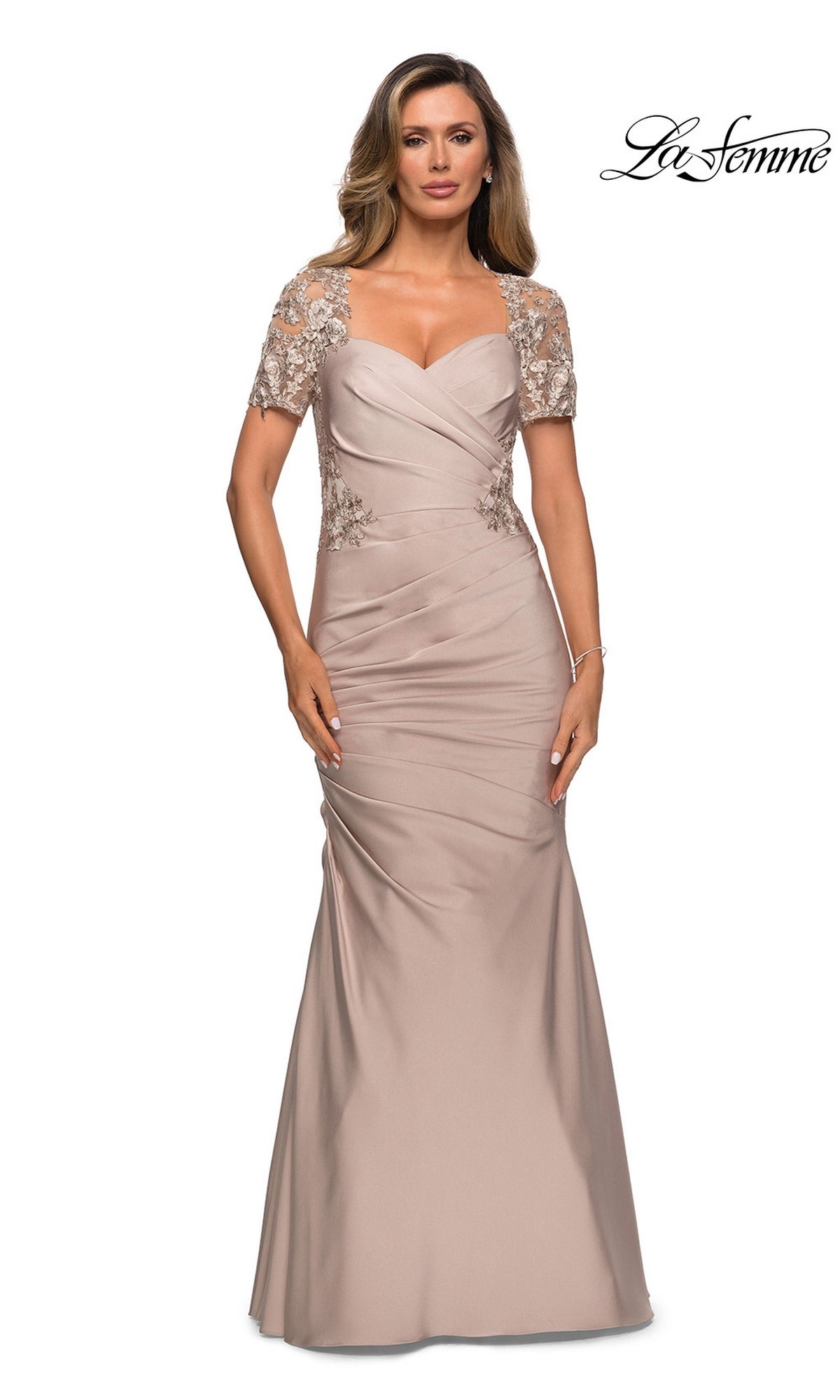 A woman poses against a white background wearing the La Femme 27989 long formal dress, featuring a floor-length taupe satin-jersey fabric. The gown has short embellished sleeves, a ruched bodice, and subtle lace applique details that highlight its elegant trumpet silhouette. The “La Femme” logo appears in the top right corner.