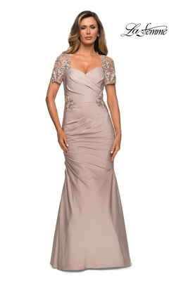 A woman poses against a white background wearing the La Femme 27989 long formal dress, featuring a floor-length taupe satin-jersey fabric. The gown has short embellished sleeves, a ruched bodice, and subtle lace applique details that highlight its elegant trumpet silhouette. The “La Femme” logo appears in the top right corner.