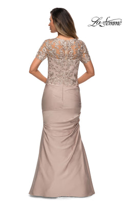 A woman stands with her back to the camera, showcasing the graceful trumpet silhouette of the Long Formal Dress: La Femme 27989. The blush-colored gown features short lace sleeves and intricate embroidery on the bodice, highlighting her figure. The “La Femme” logo appears in the top right corner.