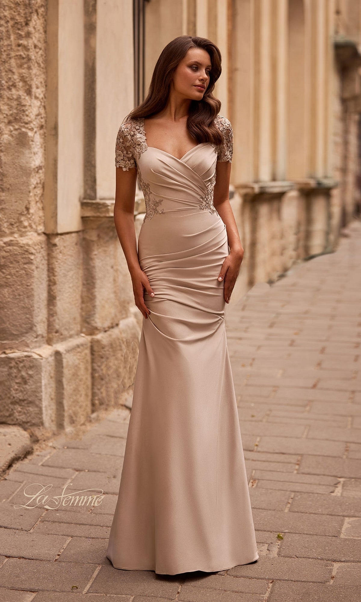 A woman wearing the Long Formal Dress: La Femme 27989—a fitted, floor-length beige evening gown featuring lace cap sleeves and delicate lace appliqué—stands on a stone-paved sidewalk beside a textured building wall.