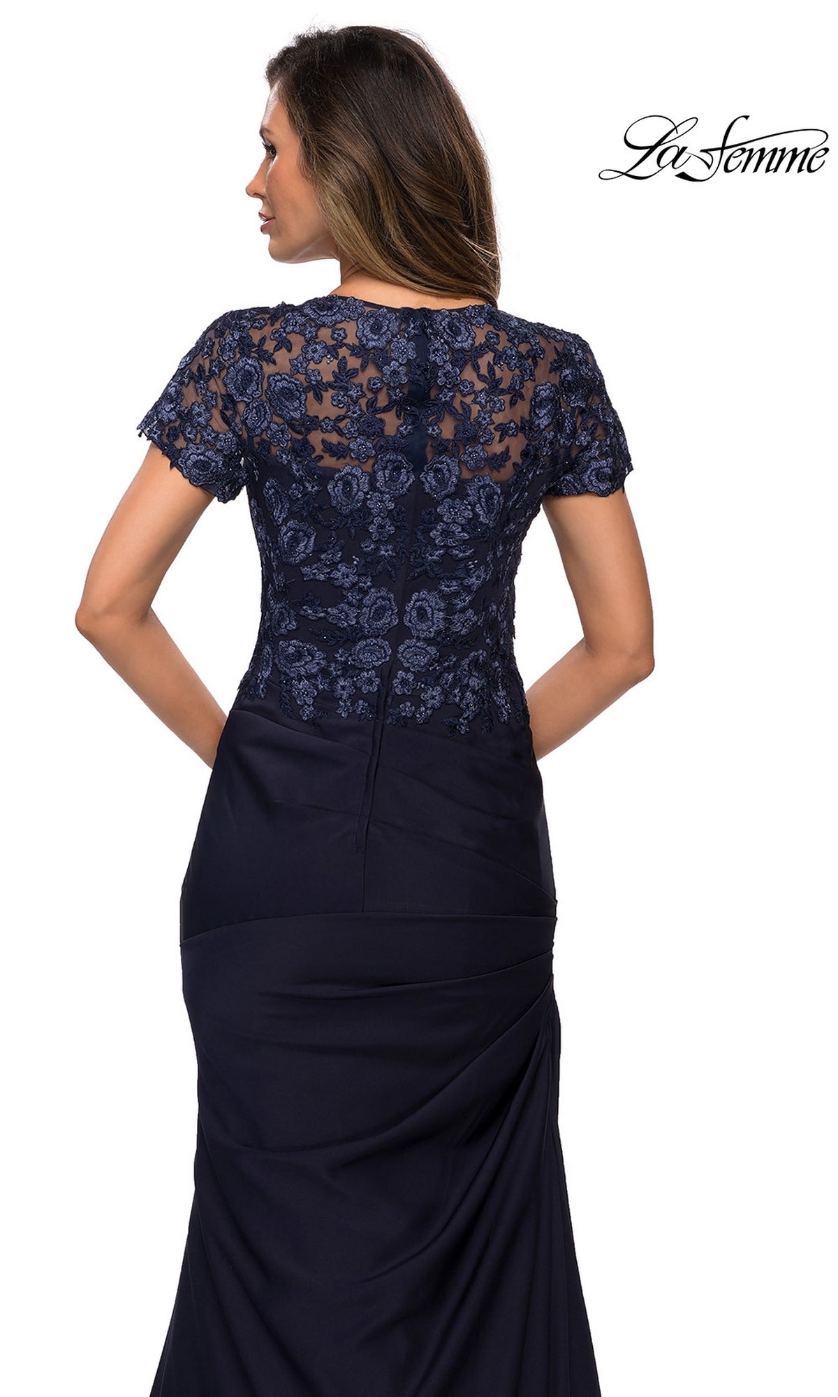 A woman is seen from behind wearing the Long Formal Dress: La Femme 27989, a navy evening gown featuring intricate lace applique detailing on the upper back and sleeves. The fitted skirt with side pleats accentuates its elegant trumpet silhouette. The La Femme logo appears in the upper right corner.
