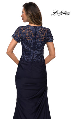 A woman is seen from behind wearing the Long Formal Dress: La Femme 27989, a navy evening gown featuring intricate lace applique detailing on the upper back and sleeves. The fitted skirt with side pleats accentuates its elegant trumpet silhouette. The La Femme logo appears in the upper right corner.