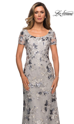 A woman wears the Long Formal Dress: La Femme 27991, a floor-length silver gown with a scoop neckline, short sleeves, and floral embroidery with sequin accents.