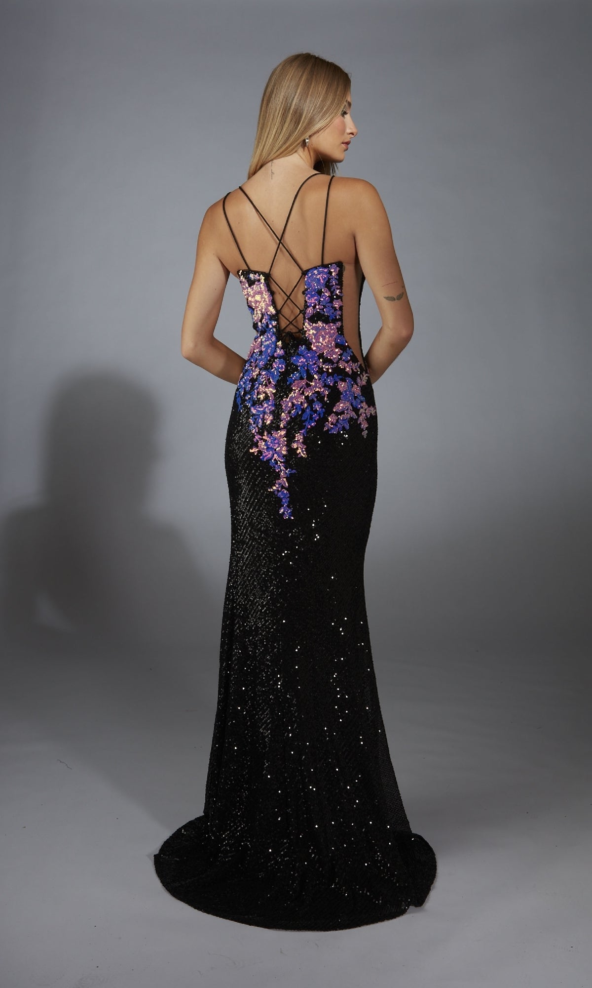 A woman faces away, wearing the Long Formal Dress: Aerie Couture 279—a black sequin gown with purple floral motifs and elegant crisscross back straps.