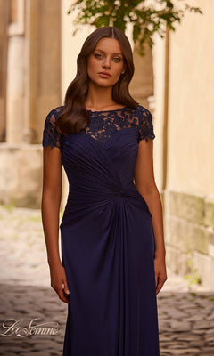 A woman stands outdoors in the Long Formal Dress: La Femme 28029, featuring a navy blue lace bodice and A-line skirt amid stone buildings and a cobblestone path.