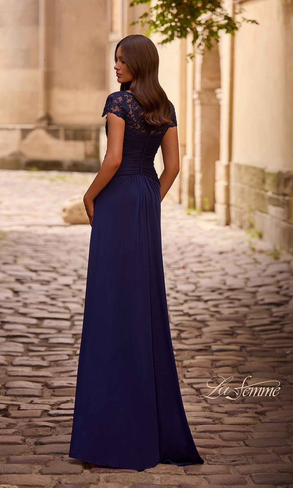 A woman in the Long Formal Dress: La Femme 28029, a navy floor-length gown with lace accents, stands on a cobblestone path facing away from the camera.