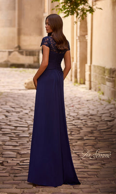 A woman in the Long Formal Dress: La Femme 28029, a navy floor-length gown with lace accents, stands on a cobblestone path facing away from the camera.