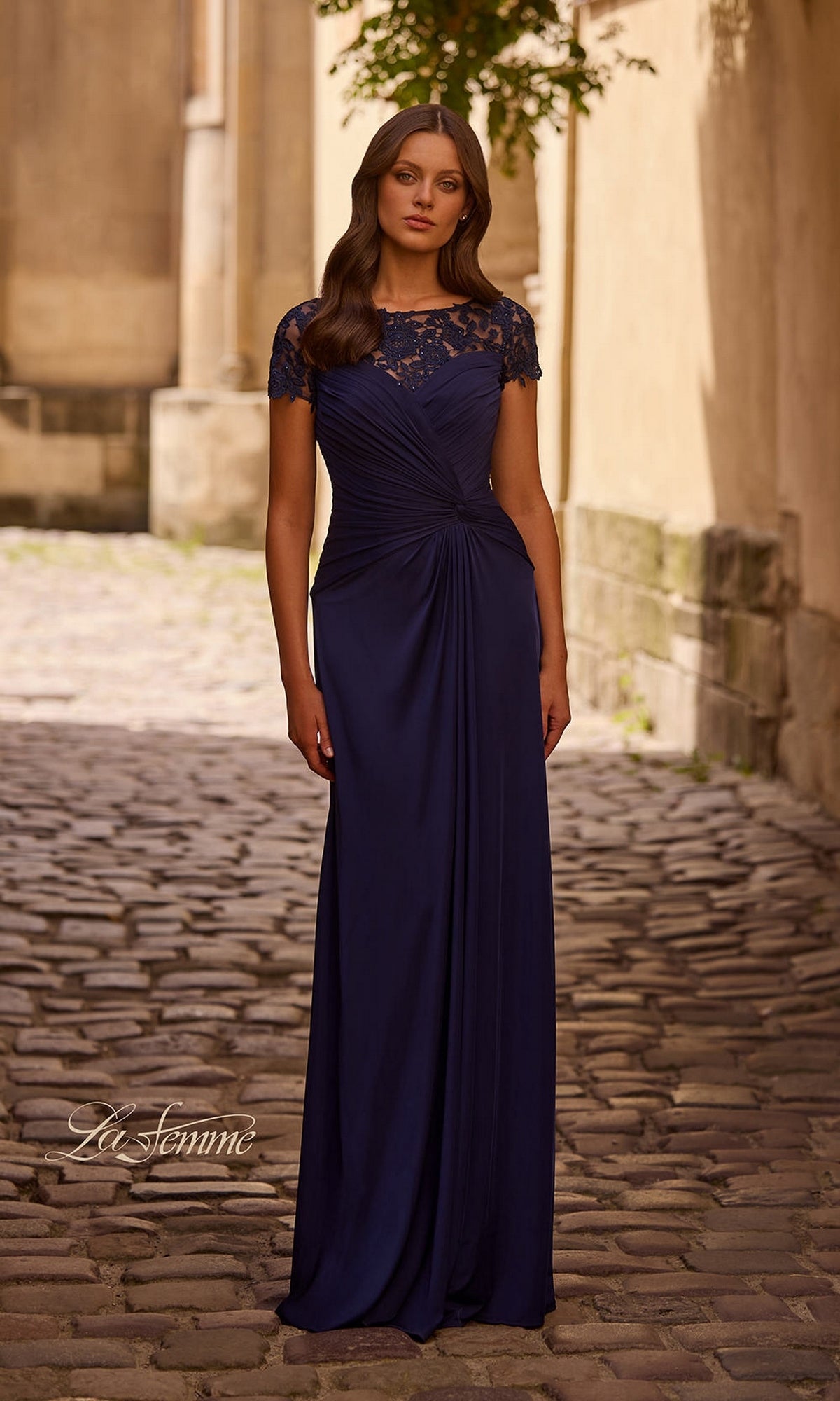 A woman stands on a cobblestone street in the Long Formal Dress: La Femme 28029, navy, featuring lace cap sleeves, a gathered waist, and an elegant A-line skirt.