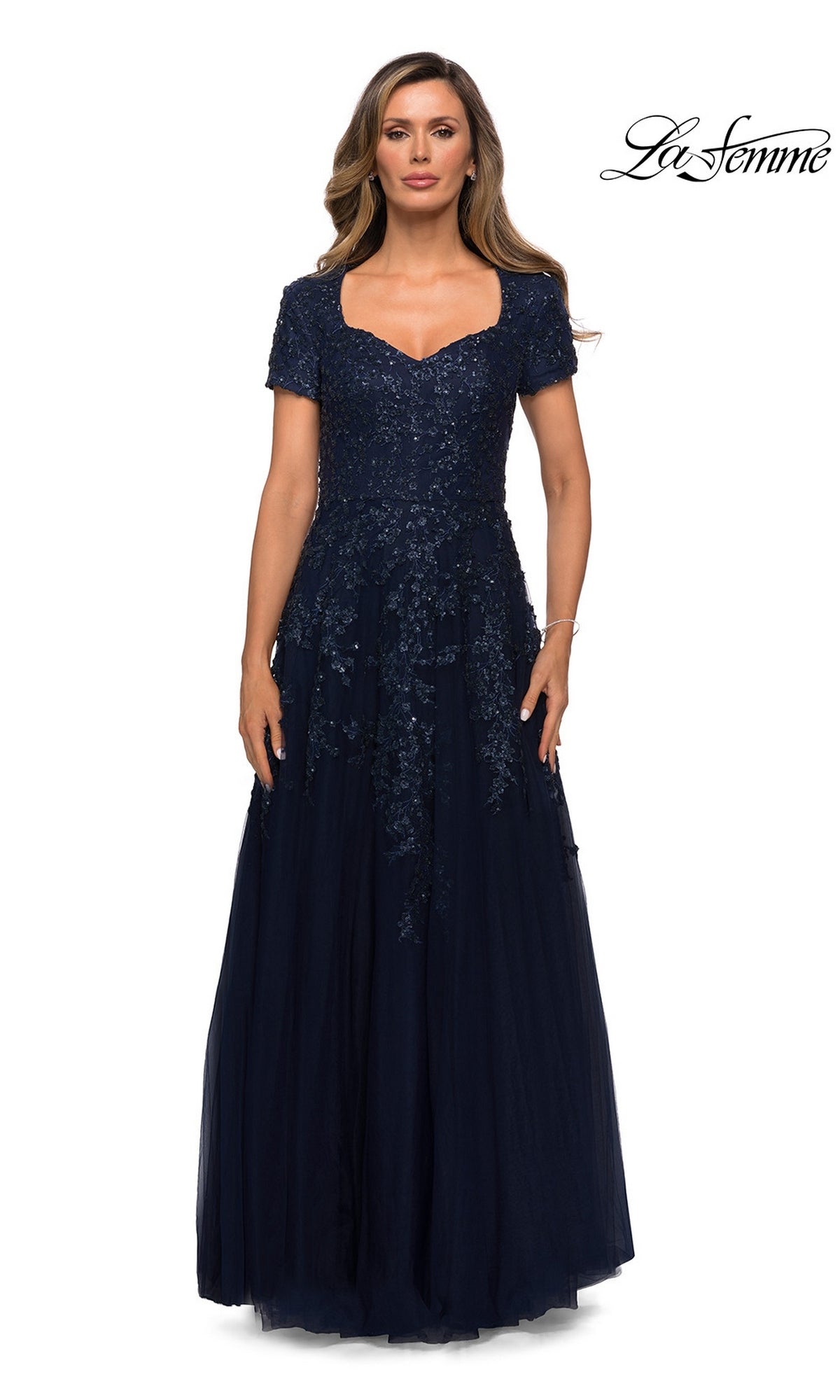 A woman wears a navy La Femme 28037 floor-length A-line gown featuring a sweetheart neckline, short sleeves, and a bodice with sequined floral appliqué.