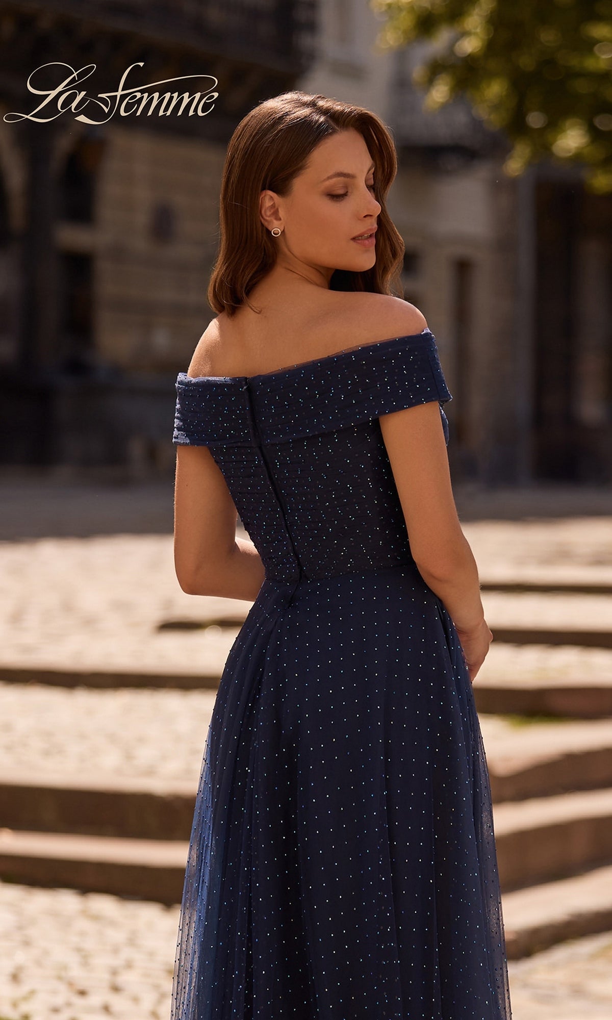 A woman faces away outdoors in a navy blue, polka-dotted Long Formal Dress: La Femme 28051. Stone steps and a building are behind her; logo appears in the top left.