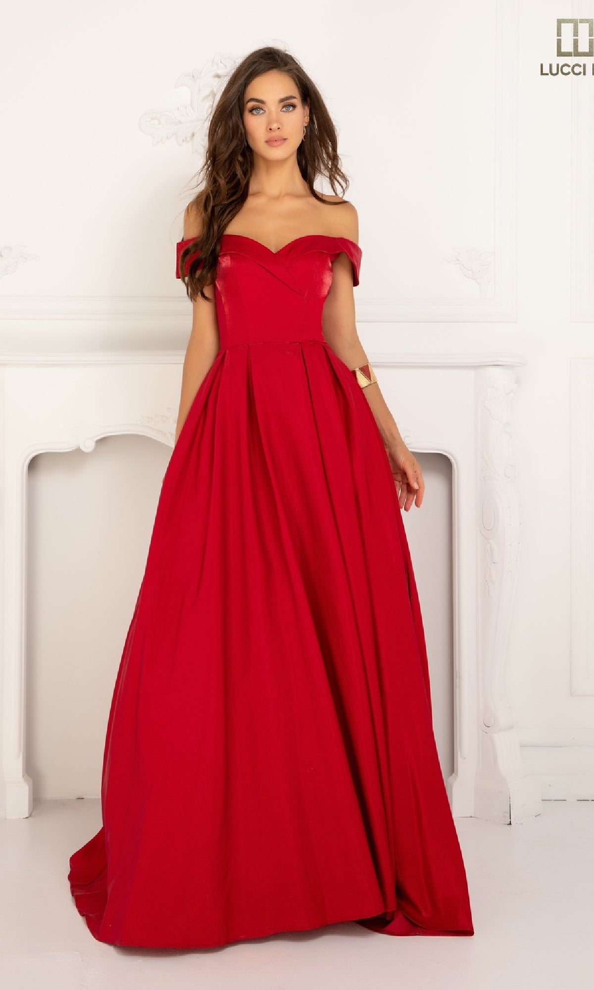 A woman stands indoors wearing the Lucci Lu 28063, an off-the-shoulder satin prom dress featuring a fitted bodice and a full, elegant floor-length red skirt.