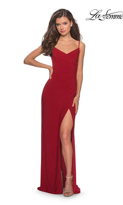 A woman in the La Femme 28079 long prom dress, showcasing a deep red sleeveless jersey design with a striking thigh-high slit and chic faux wrap silhouette, poses with one hand on her hip and pairs the look with strappy heels.