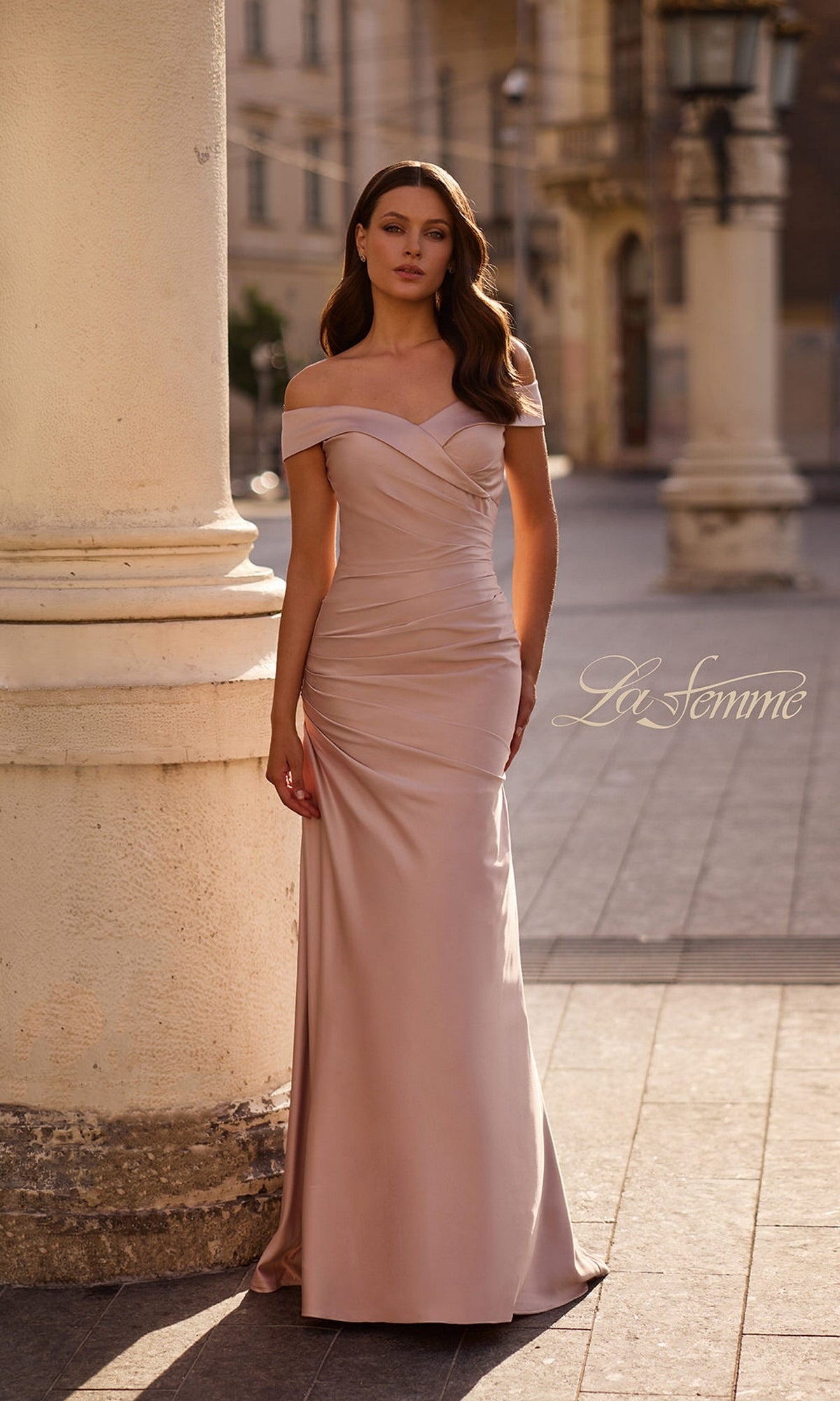 A woman in a pink champagne off-the-shoulder gown, the Long Formal Dress: La Femme 28103—ideal for a mother-of-the-bride—stands outdoors near columns, with the “La Femme” logo displayed on the image.