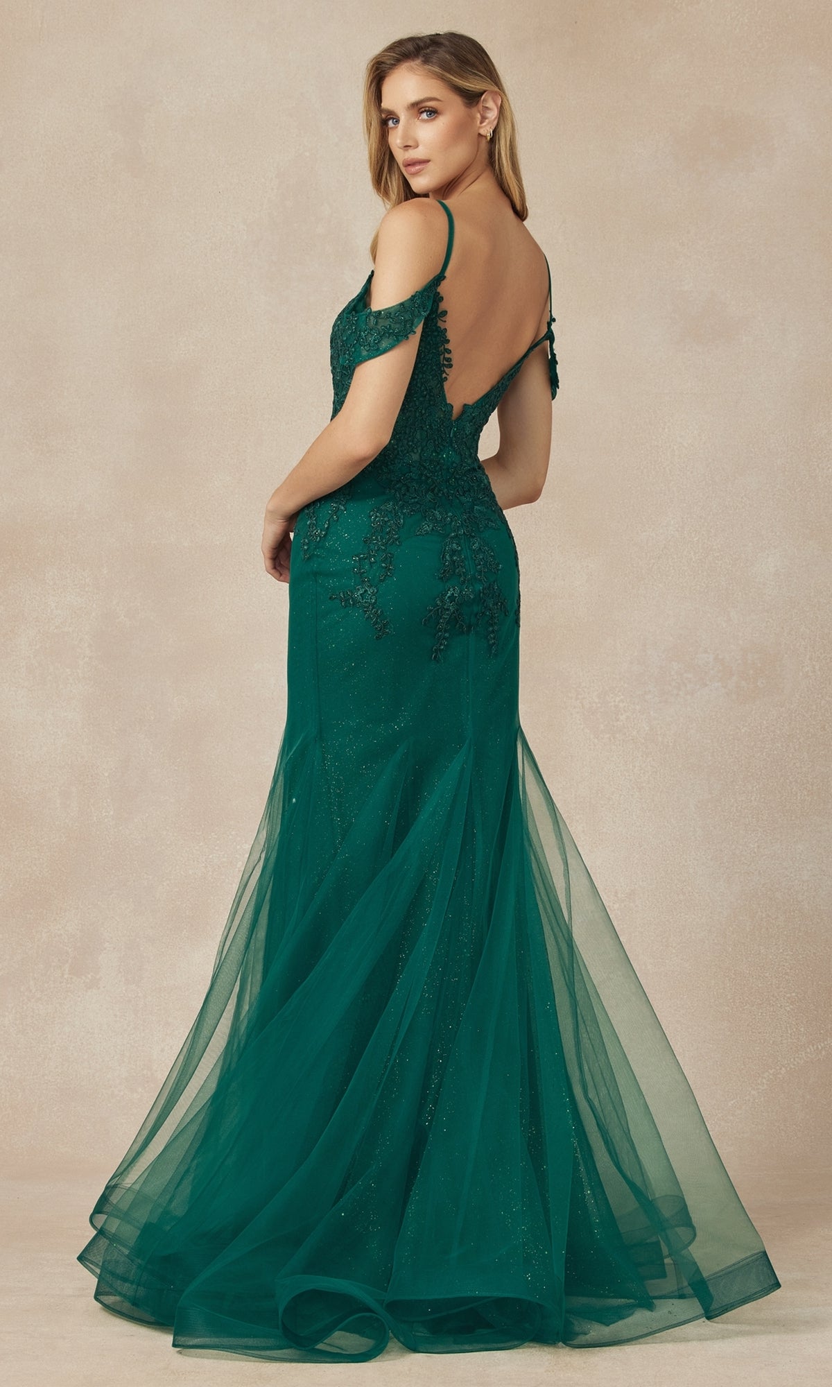 A woman wears the Juliet 281 Long Glitter Mermaid Prom Dress in emerald green, featuring off-the-shoulder lace details and a sheer tulle skirt, as she stands facing away and looks over her shoulder.