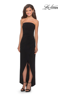 Woman in a black Long Prom Dress: La Femme 28204 with a strapless ruched bodice and front slit, paired with black heels, smiling against a white background.