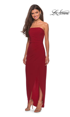 A woman wears the Long Prom Dress: La Femme 28204 in red with a ruched bodice and sheath skirt, beige heels, and a white background featuring the “La Femme” logo.