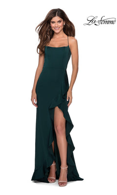 Woman wearing a dark green La Femme Side-Ruffle Long Prom Dress 28294 with an open back, spaghetti straps, high ruffled slit, and strappy nude heels, standing and smiling against a white background.