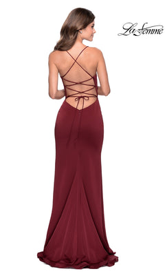 A woman wearing a floor-length, fitted La Femme Side-Ruffle Long Prom Dress 28294 in burgundy with open back and crisscross lace-up straps stands against a white background.