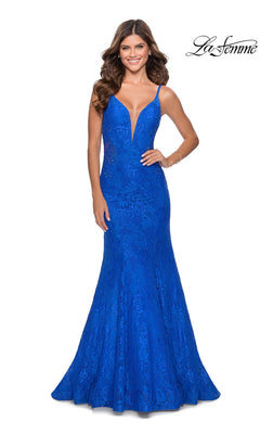 A woman models the La Femme Backless Long Lace Prom Dress 28355 in royal blue, featuring a deep V-neckline, fitted bodice, and flared mermaid skirt, standing against a white backdrop.
