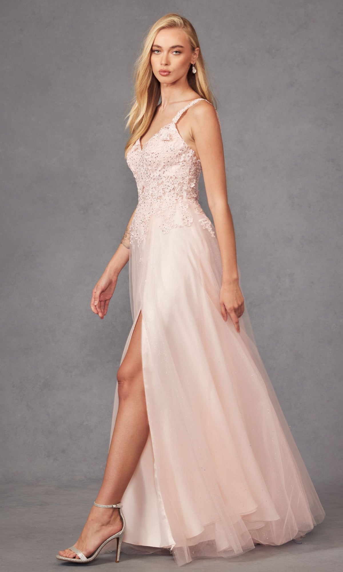A woman poses against a gray backdrop wearing the Long Prom Dress 283 by Juliet—a pale pink, sleeveless gown featuring lace embroidery, bead embellishments, and a thigh-high slit.