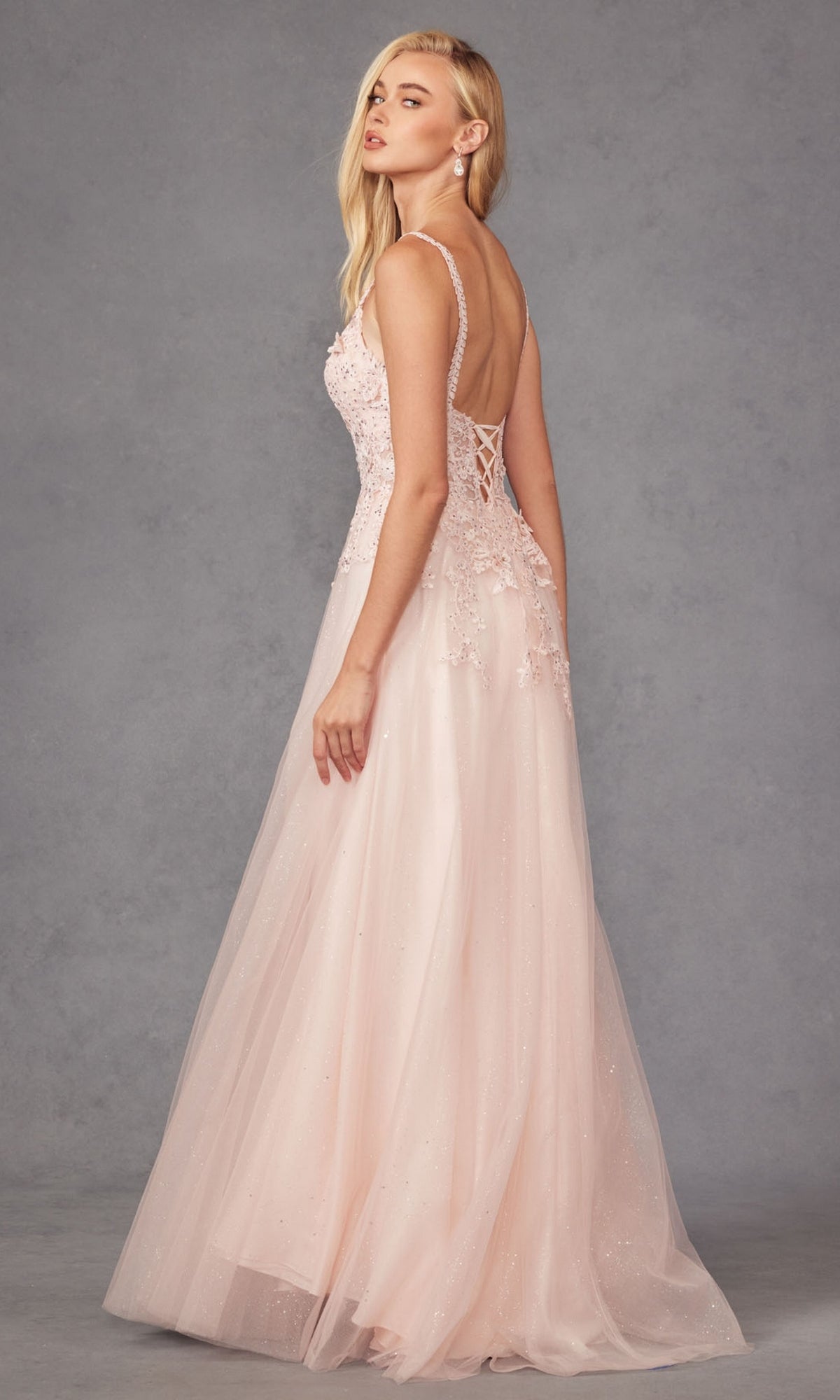 A woman wears the Long Prom Dress 283 by Juliet—a light pink, sleeveless, lace-embroidered A-line gown with a v-neckline and lace-up back—standing against a plain gray background.