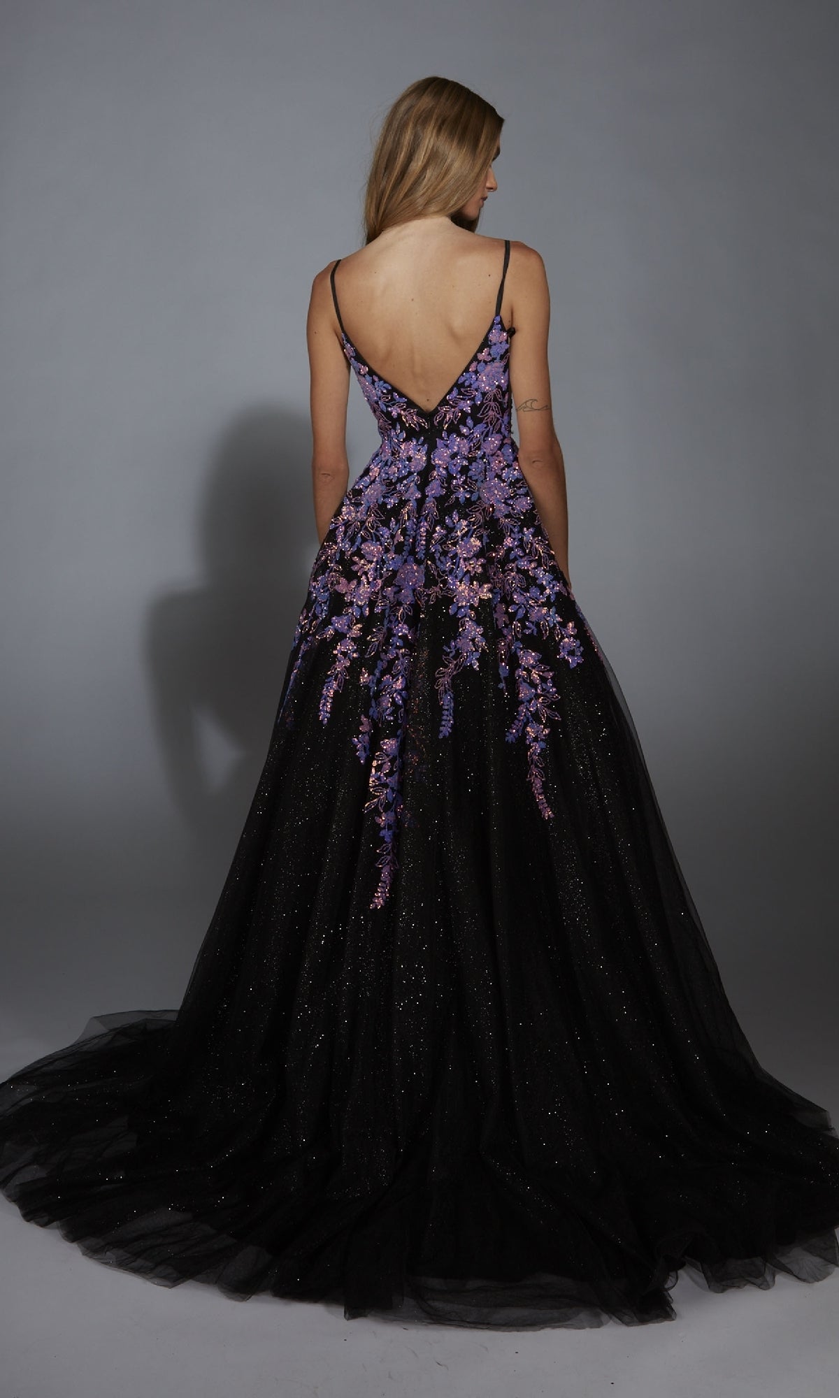 A woman faces away in the Aerie Couture 283, a long formal dress with a deep V-back and embellished floral details flowing down its elegant glitter tulle A-line skirt.